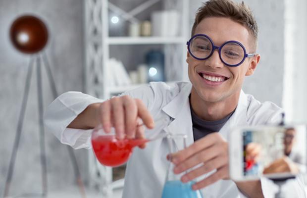 young man chemist pouring red liquid from one beaker to another, smiling